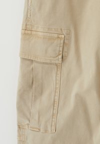Beige puuvillakankaiset cargo-shortsit, joissa on suuri sivutasku, ommellut korostukset ja sileä, hieman ryppyinen pinta.