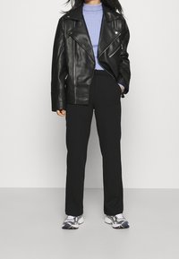 Osoba w czarnej skórzanej kurtce motocyklowej, jasnoniebieskim prążkowanym golfie, czarnych prostych spodniach i szaro-białych sneakersach stoi na szarym podłodze.