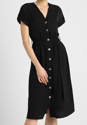 Femme portant une robe noire à boutons, manches courtes, col en V et ceinture nouée à la taille, debout devant un fond uni.