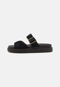 Sandale noire en daim avec un bout ouvert et une large sangle, ornée d'une boucle dorée sur une semelle noire contournée. Design plat avec des détails minimalistes.