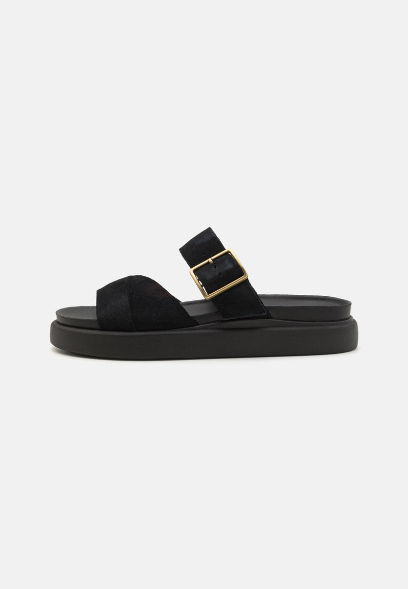 Sandale noire en daim avec un bout ouvert et une large sangle, ornée d'une boucle dorée sur une semelle noire contournée. Design plat avec des détails minimalistes.