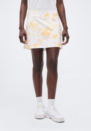 Άτομο που φοράει λευκή και κίτρινη φούστα με μοτίβο tie-dye, λευκές κάλτσες και λευκά αθλητικά παπούτσια, στέκεται μπροστά σε απλό λευκό φόντο.