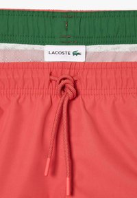 Pantaloni da bagno rossi con una cintura verde, dotati di un laccio frontale e un'etichetta del logo Lacoste sotto un rivestimento in rete.