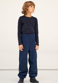 Marineblaue wasserdichte Hose mit hoher Taille, elastischen Bündchen und verstärkten Knieflicken. Kombiniert mit einem figurbetonten langärmeligen dunklen Oberteil.