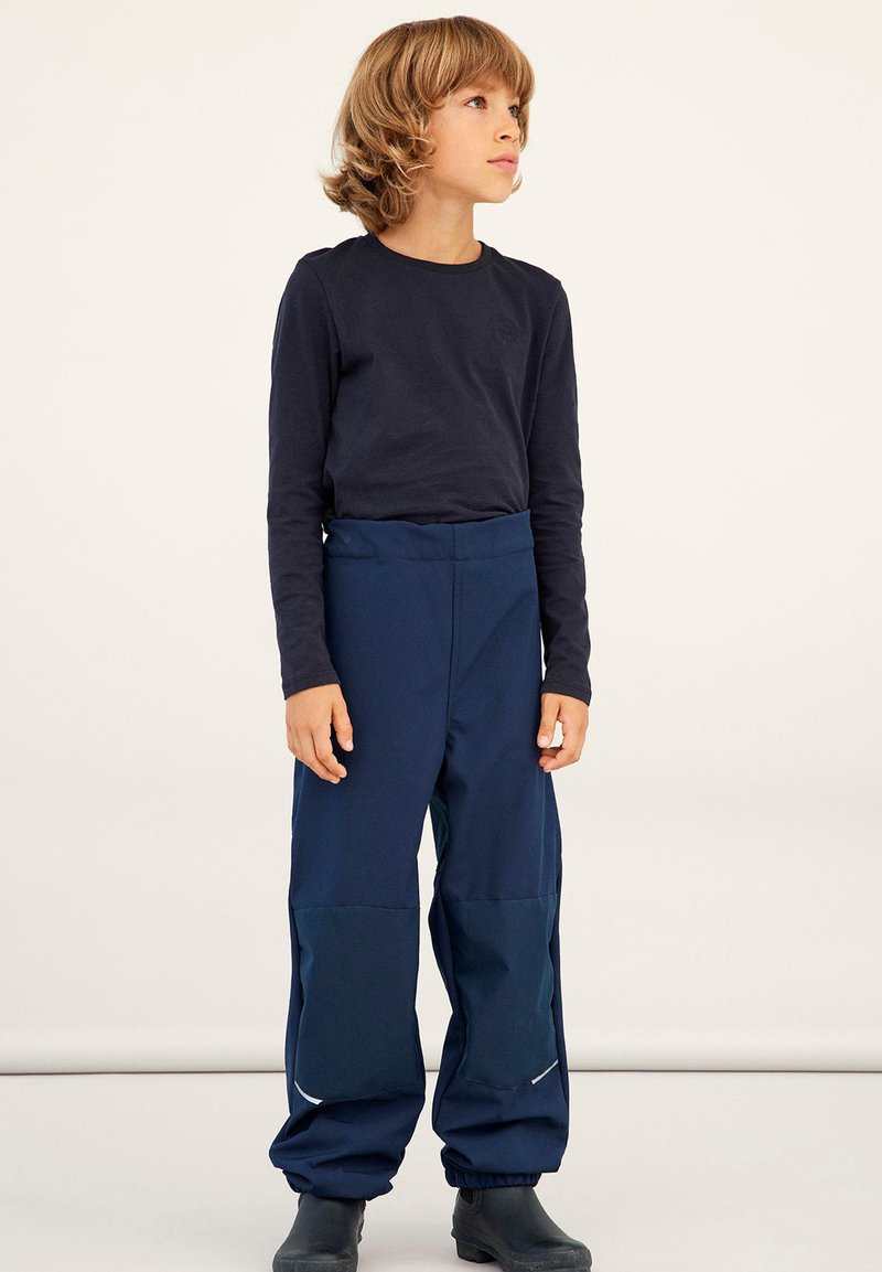 Marineblaue wasserdichte Hose mit hoher Taille, elastischen Bündchen und verstärkten Knieflicken. Kombiniert mit einem figurbetonten langärmeligen dunklen Oberteil.