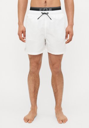 Shorts de bain blancs avec une taille élastique noire comportant un logo, fermeture à cordon, et poignets repliés. Le matériau semble léger et lisse.