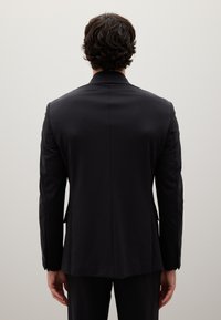 Blazer sartoriale nero con una texture liscia, spacco singolo sul retro e leggero lucido; presenta colletto a punte e polsini abbottonati.