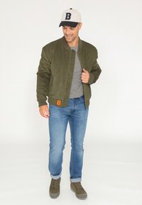 Blouson aviateur vert olive avec des poignets et une taille côtelés, associé à un t-shirt gris et un jean bleu, complété par une casquette beige et noir.