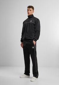 Costum de trening negru cu detalii albe, compus dintr-o jachetă cu fermoar și pantaloni cu talie elastică. Include logo pe jachetă și pantaloni. Pantofi sport albi.