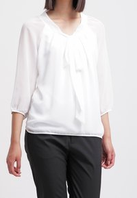 Woman wears a white sheer blouse with a front tie and three-quarter sleeves, paired with black fitted trousers against a plain background.