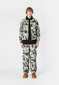 Conjunto de chaqueta y pantalones de camuflaje en tonos de verde, beige y negro, con cierre de cremallera, bolsillos y textura de tela suave. Calzado marrón.