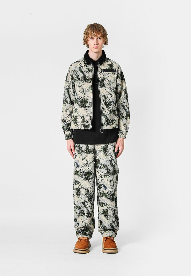 Conjunto de chaqueta y pantalones de camuflaje en tonos de verde, beige y negro, con cierre de cremallera, bolsillos y textura de tela suave. Calzado marrón.