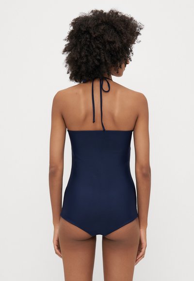 Sötétkék egyrészes fürdőruha, amely nyakba köthető pánttal, egyszínű textúrát és a testet kiemelő elegáns dizájnt kínál.