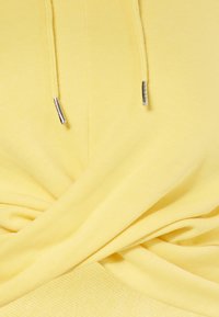 Sudadera amarilla con un diseño anudado en la parte frontal. Presenta cordones delgados con puntas metálicas y una textura de algodón suave.