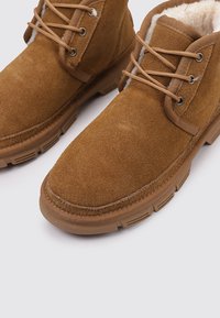 Botas de ante marrón con cordones, suelas de goma gruesa y un suave forro de lana blanca, mostradas una al lado de la otra sobre un fondo blanco.