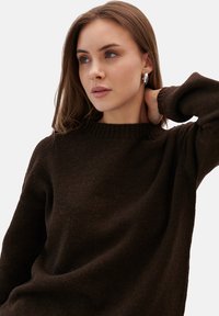 Brązowy sweter z dzianiny z prążkowanym dekoltem, miękką teksturą i długimi rękawami. Modelka ma rękę opartą na szyi.