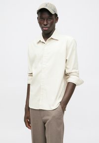 Chemise beige à manches longues, en tissu léger texturé, associée à un pantalon marron clair et une casquette beige. Design simple et épuré.