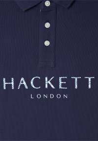 Námořnická modrá polo košile s texturovanou látkou, která má tříknoflíkovou légu a potištěné logo "HACKETT LONDON" v světle modrých akcentu.