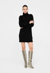 Abito in maglia nera con collo alto, dotato di due tasche frontali. Abbinato a stivali a stampa serpente con tacco dorato fino al ginocchio.