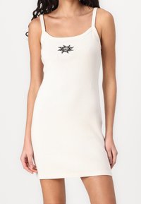 Femme portant une mini-robe sans manches, de couleur claire, avec de fines bretelles et un petit motif noir de soleil sur la poitrine.