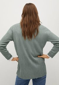 Maglietta verde a coste con una vestibilità morbida, maniche lunghe e orlo asimmetrico. Presenta un accento bianco sulla manica che spunta da sotto.