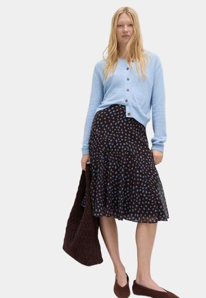 Frau trägt hellblauen, geknöpften Cardigan, schwarzen Rock mit blauen Punkten, hält eine große braune strukturierte Tasche und trägt braune flache Schuhe.
