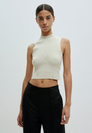 Geribbelde beige crop top met een hoge hals en V-vormige detail, gecombineerd met zwarte high-waisted broek. Glad textuur en getailleerd ontwerp.