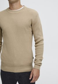 Beige gebreide trui met een textuur en wafelpatroon, en geribbelde manchetten en zoom. Gecombineerd met donkere spijkerbroeken voor een casual look.