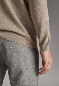 Zbliżenie na prawą rękę osoby spoczywającą obok ciała, ubraną w beżowy sweter wykonany na drutach i szare wełniane spodnie na jednolitym tle.