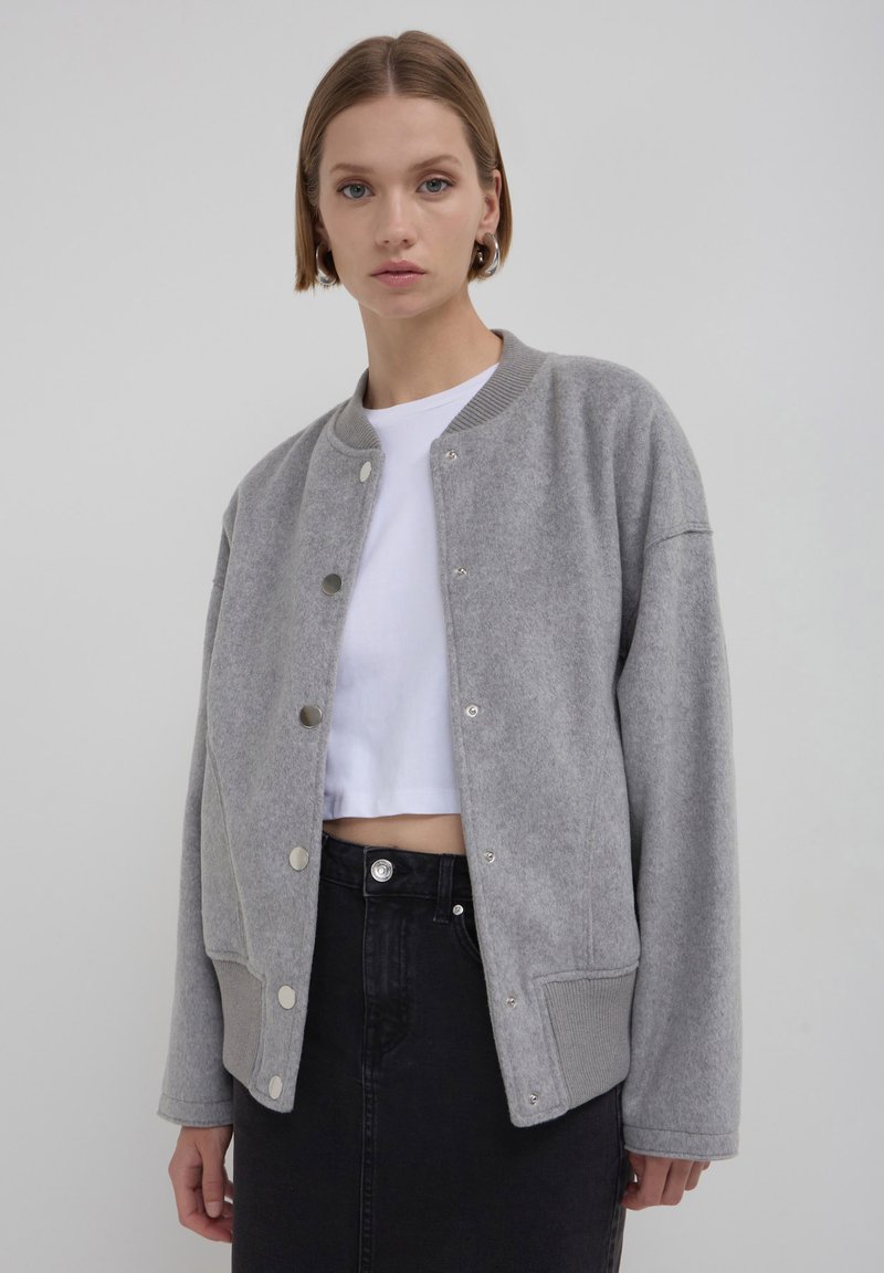 Frau mit kurzen Haaren, die silberne Creolen, eine graue, geknöpfte Jacke, ein weißes Cropped-Top und einen hochgeschnittenen schwarzen Rock trägt, vor einem grauen Hintergrund.