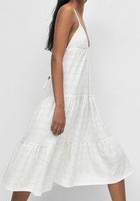 Bílé bavlněné šaty s vrstveným designem, které mají hluboký výstřih do V, tenké ramínka, výšivku a boční zavázání s dekorativním třásní.