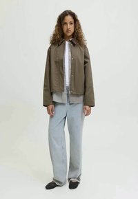 Bruine cropped jacket met een drukknopen-sluiting aan de voorkant over een zwart-witte geruite blouse, gecombineerd met lichtblauwe rechtgesneden jeans en zwarte ballerina's.