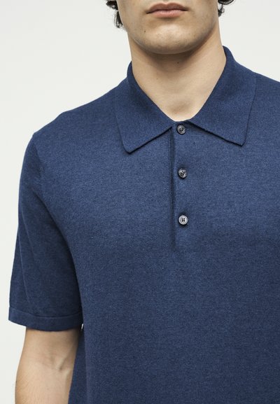 Άνδρας που φοράει μπλούζα πόλο με κοντά μανίκια, σε ναυτικό μπλε χρώμα, με γιακά και τρία κουμπιά, εμφανιζόμενος από τους ώμους μέχρι το μέσο του στήθους.