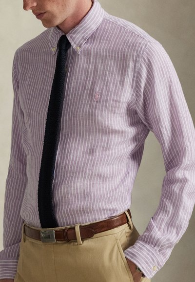 Πουκάμισο με κουμπιά σε ανοιχτό μωβ χρώμα με λευκές ρίγες, διαθέτει μικρό λογότυπο στο στήθος, συνδυασμένο με υφασμάτινη μαύρη γραβάτα και χακί παντελόνι.