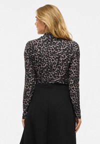 Femme blonde de dos portant un haut à manches longues en imprimé léopard rentré dans une jupe noire taille haute, devant un fond blanc.