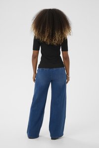 Femeie cu păr creț, îmbrăcată cu un top negru cu mâneci scurte și blugi largi de culoare albastră, stând cu fața îndreptată spre exterior pe un fundal alb.