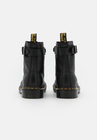 Coppia di stivaletti alla caviglia in pelle nera con cuciture gialle, linguette con la scritta "WITH BOUNCING SOLES" e cinturini con fibbia visti da dietro.