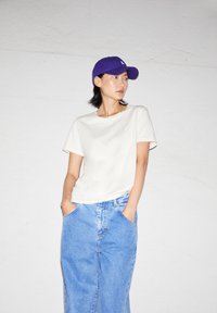 Weißes T-Shirt mit kurzen Ärmeln und hellblauen, locker sitzenden Jeans. Eine lila Mütze mit einem Logo sitzt auf dem Kopf. Die Textur wirkt glatt und lässig.