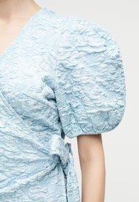 Φόρεμα από υφασμάτινο ύφασμα σε ανοιχτό γαλάζιο χρώμα με φουσκωτά κοντά μανίκια και λεπτομέρεια τύλιγμα γύρω από τη μέση, πάνω σε άτομο με ορατό γυμνό χέρι.