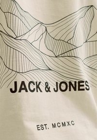Kreem värvi T-särk musta joonistusega lainelistest mägedest. Tekst "JACK & JONES" ja "EST. MCMXC" paksus kirjas. Pehme puuvilla tekstuur.