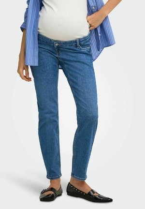 Gravid person iført blå graviditetsjeans, hvid top, blåstribet skjorte og sorte flade sko med metalnitter.