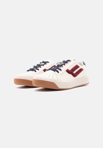 Baskets blanches basses avec semelles marron, lacets bleu marine et logo latéral texturé rouge foncé, présentées en angle sur un fond uni clair.