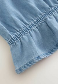 Tissu en denim bleu clair avec un détail de volant froncé à l'ourlet. Texture douce et couture bien définie sont visibles.