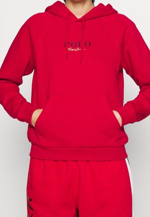 Henkilö pukeutuneena punaiseen hupulliseen paitaan, jossa on etutaskut ja rintakehällä mustavalkoinen "POLO Ralph Lauren" -logo, yhdistettynä punaisiin housuihin.