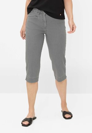Kvinde iført grå cropped bukser, sort ærmeløs top og sorte åbne slip-on sandaler mod en hvid baggrund.