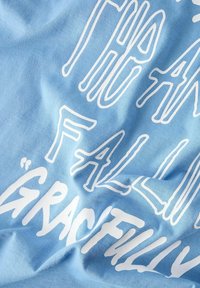 Ljusblå bomullströja med vit text som står "DEN DÄR FALLANDE GRACISKT." Slät textur med lätt veck och en avslappnad utseende.