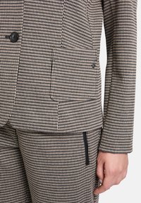 Costume texturé en tissu à motif pied-de-poule beige et noir. Il comporte deux poches avant et une fermeture à un bouton.