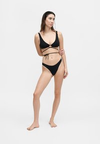 Γυναίκα που στέκεται με τα χέρια σταυρωμένα, φοράει μαύρο μπικίνι με κορδόνια που τυλίγονται γύρω, κοιτάζει ελαφρώς στο πλάι σε απλό φωτεινό φόντο.