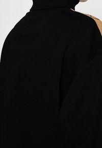 Sudadera negra con cuello alto y un panel en contraste de color beige en el hombro, con una textura suave y un ajuste relajado.