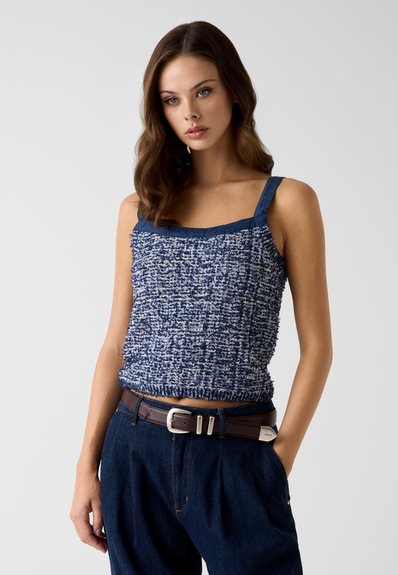 Femme aux cheveux bruns ondulés portant un top sans manches texturé bleu et blanc et un pantalon taille haute bleu foncé avec une ceinture marron, debout.